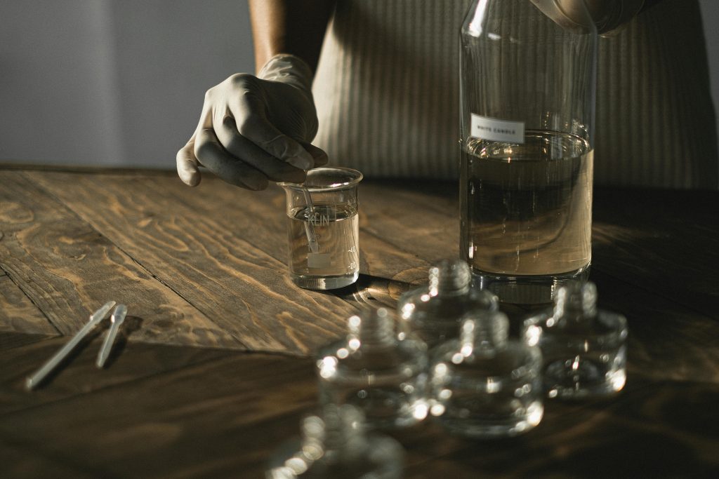 Auf dem Bild sind mehrere kleine Glasfläschchen mit klaren und farbigen Duftessenzen zu sehen. Eine Hand hält eine Pipette und gibt präzise Tropfen einer Substanz in ein Mischglas. Der Tisch ist mit Laborzubehör ausgestattet, was den Eindruck eines professionellen Parfumherstellungsprozesses vermittelt. Es entsteht der Eindruck von Konzentration, Präzision und der handwerklichen Kunst, aus einzelnen Komponenten eine harmonische Duftkomposition zu erschaffen.