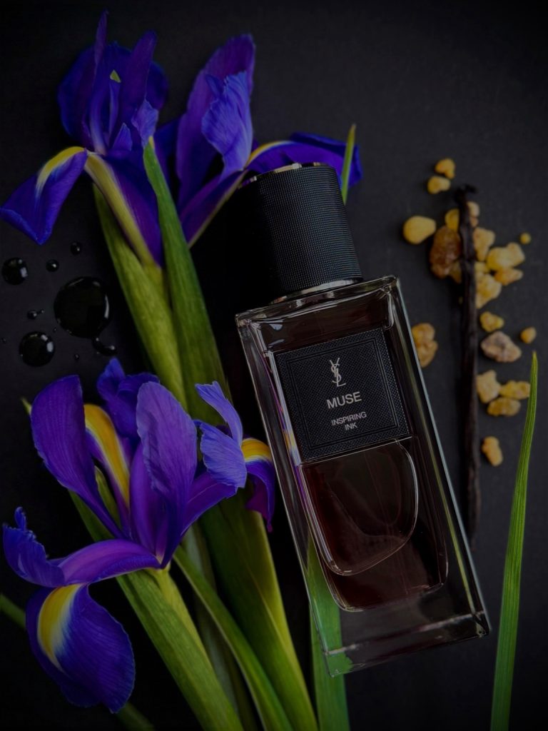 Der elegante Glasflakon von Yves Saint Laurent Muse liegt vor intensiv violetten Irisblüten. Neben ihm sind goldene Harzstücke und eine dunkle Vanilleschote arrangiert. Der schwarze Hintergrund und die gedämpfte Beleuchtung betonen die Tiefe der bernsteinfarbenen Flüssigkeit im Flakon. Die Komposition aus Iris, Harz und Vanille greift zentrale Duftfacetten auf und unterstreicht die dunkle, elegante Atmosphäre von Muse – Inspiring Ink.