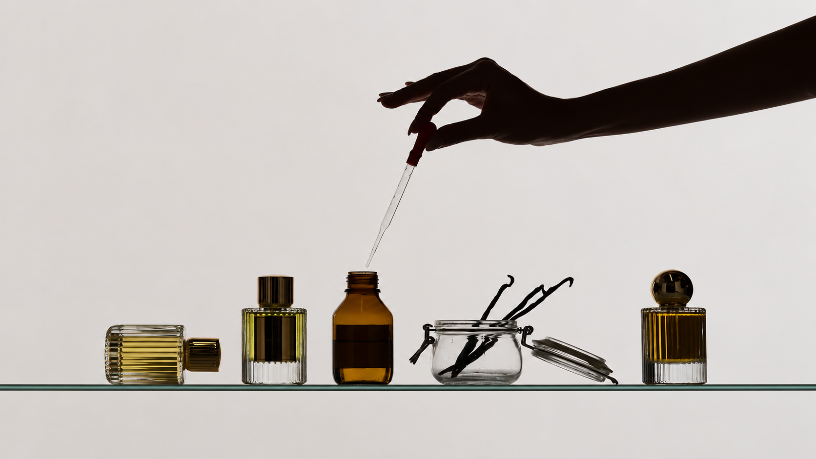 Alkohol im Parfum – Hand mit Pipette über einem Flakon, umgeben von Parfumflaschen und Vanilleschoten.